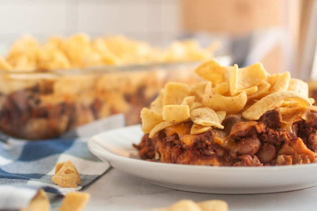 A white plate with a serving of Frito pie, topped with corn chips, is shown on a light surface.