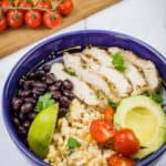 A blue bowl filled with a Street Corn Chicken Rice Bowl, featuring grilled chicken, corn salad, tomatoes, avocado, black beans, and lime.