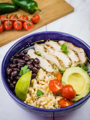A blue bowl filled with a Street Corn Chicken Rice Bowl, featuring grilled chicken, corn salad, tomatoes, avocado, black beans, and lime.