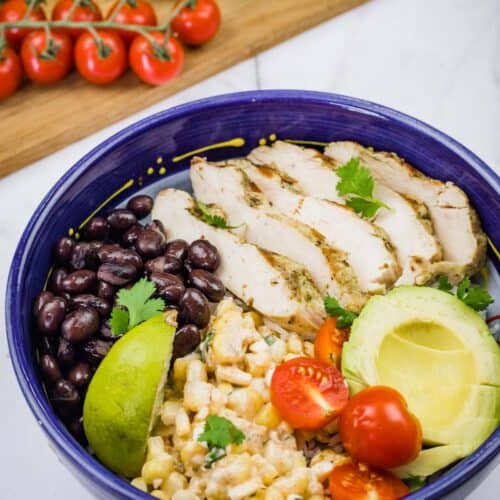 A blue bowl filled with a Street Corn Chicken Rice Bowl, featuring grilled chicken, corn salad, tomatoes, avocado, black beans, and lime.
