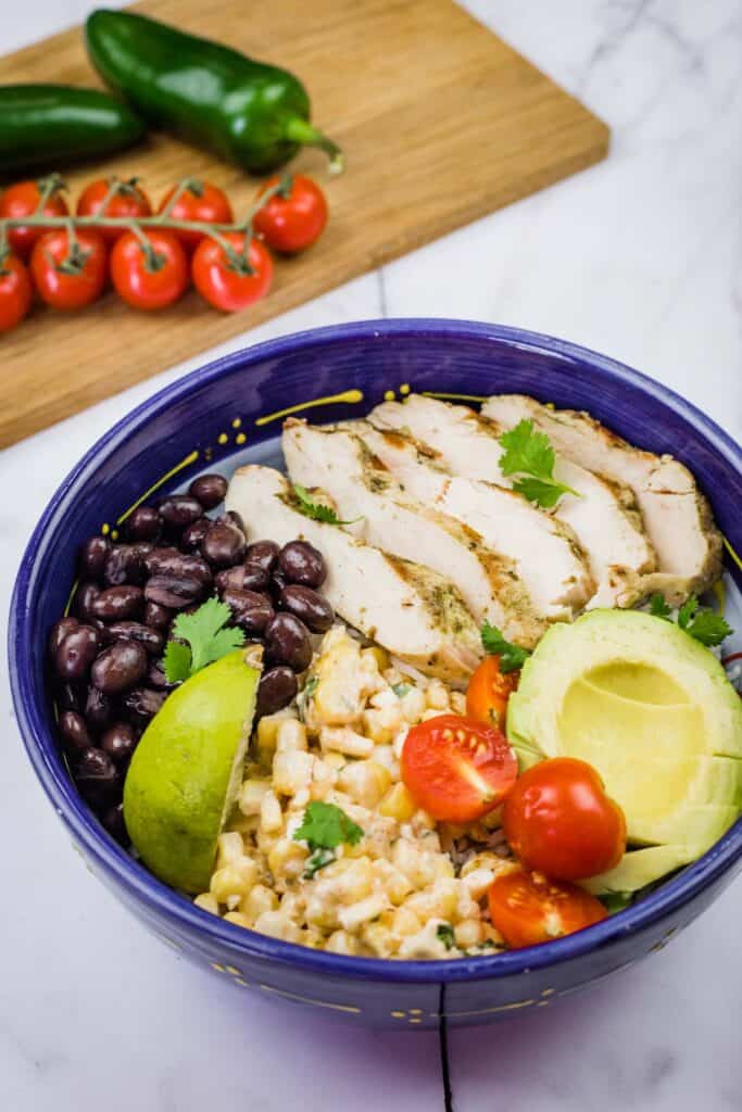 A blue bowl filled with a Street Corn Chicken Rice Bowl, featuring grilled chicken, corn salad, tomatoes, avocado, black beans, and lime.