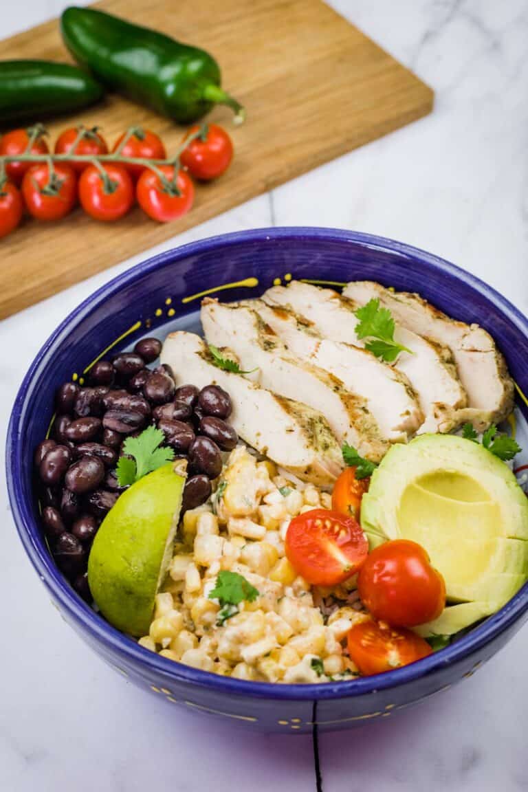 A blue bowl filled with a Street Corn Chicken Rice Bowl, featuring grilled chicken, corn salad, tomatoes, avocado, black beans, and lime.
