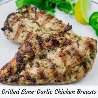 Two grilled lime-garlic chicken breasts on a plate, garnished with herbs and lime.