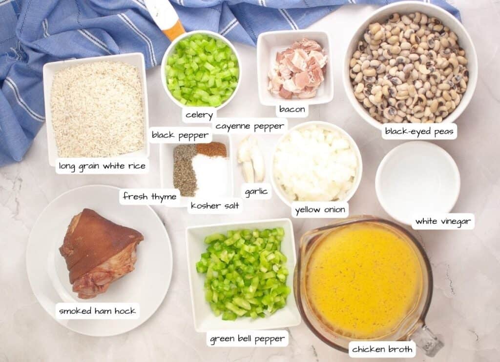 Overhead view of labeled ingredients for a dish, including rice, celery, bacon, black-eyed peas, spices, onion, vinegar, ham hock, bell pepper, garlic, and chicken broth.