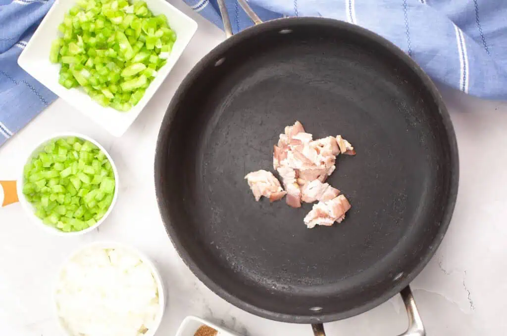 Chopped bacon pieces in a skillet with bowls of chopped celery, green bell pepper, and onion on the side.