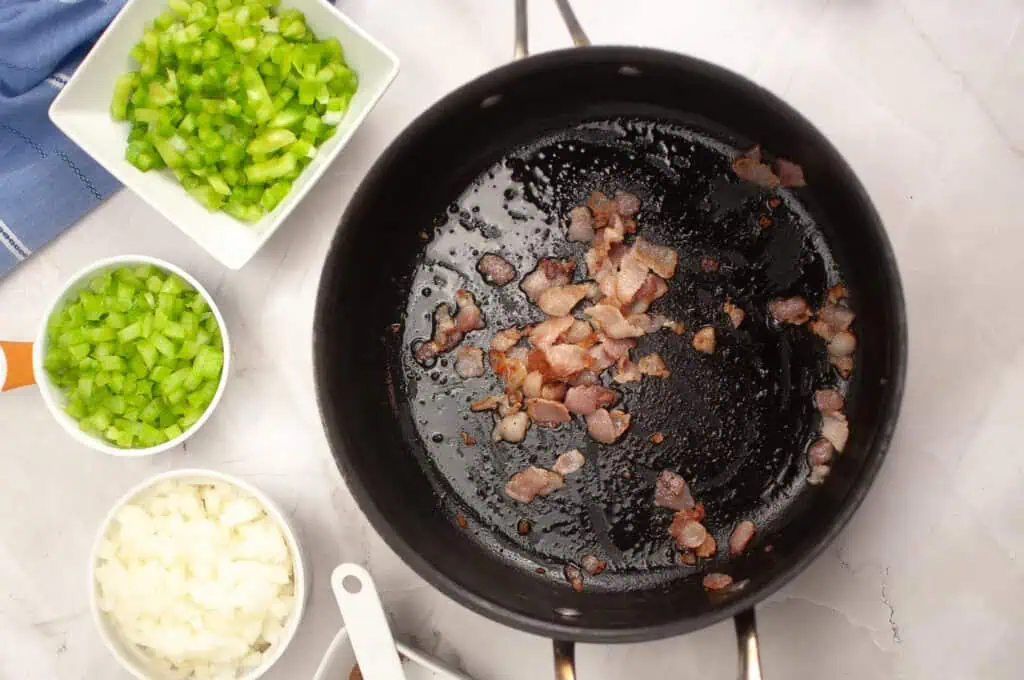 Chopped bacon cooking in a black pan, with bowls of diced celery, green bell pepper, and onion arranged nearby on a white countertop.