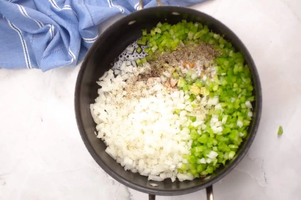 Chopped onions and green bell peppers with seasonings in a saucepan, ready to be cooked, with a blue cloth nearby on a white surface.