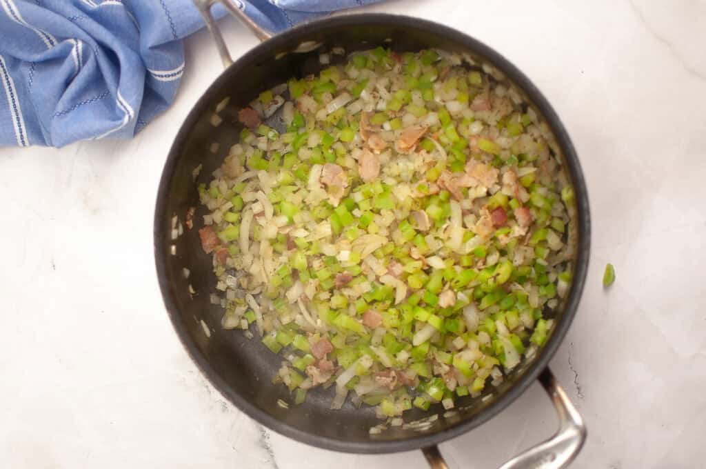 A pot containing chopped celery, onions, and bacon pieces being saut&eacute;ed on a stovetop, with a blue kitchen towel nearby.