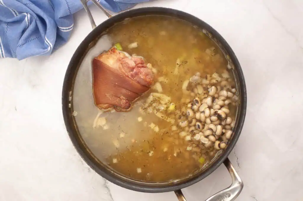 A pot of soup with a large piece of smoked meat and black-eyed peas simmering in broth on a white surface, next to a blue cloth.