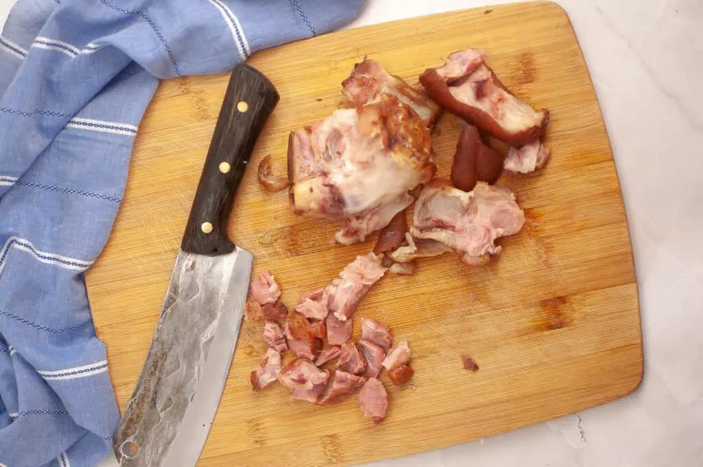 A wooden cutting board with a large knife, a blue kitchen towel, and chopped pieces of smoked ham hock.