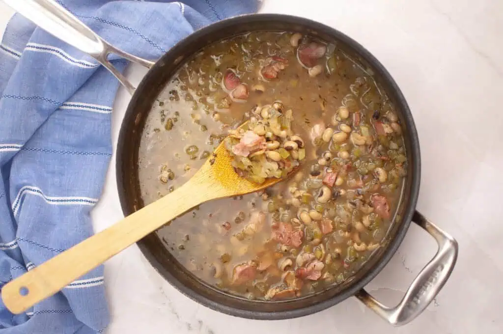 A pot of black-eyed peas with ham and vegetables simmers, with a wooden spoon resting inside; a blue kitchen towel is beside the pot on a white marble surface.