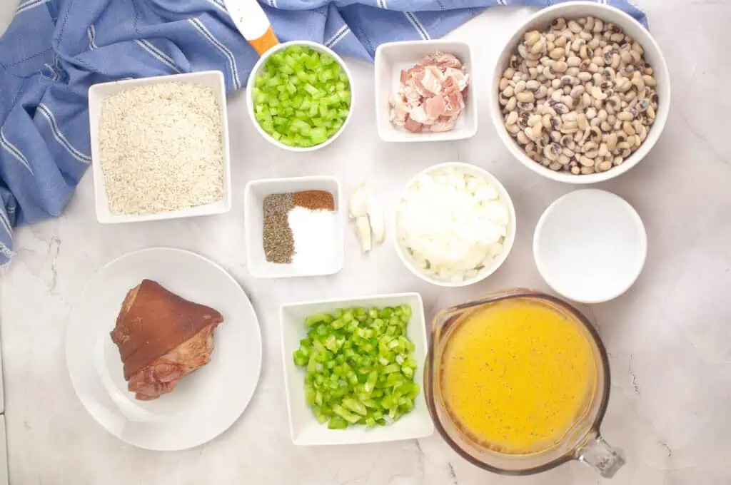 Overhead view of bowls containing rice, chopped celery, chopped onions, black-eyed peas, ham, seasonings, garlic, water, and broth on a white surface with a blue cloth.