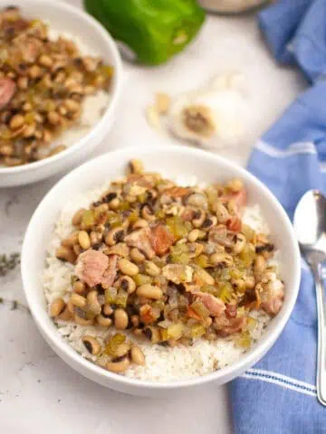 A bowl of rice topped with black-eyed peas, diced ham, and vegetables sits on a marble surface next to a spoon, a green bell pepper, garlic, and a blue napkin.