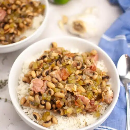 A bowl of rice topped with black-eyed peas, diced ham, and vegetables sits on a marble surface next to a spoon, a green bell pepper, garlic, and a blue napkin.