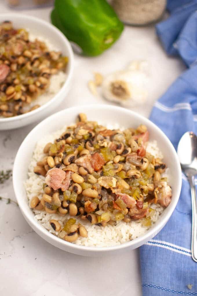 A bowl of rice topped with black-eyed peas, diced ham, and vegetables sits on a marble surface next to a spoon, a green bell pepper, garlic, and a blue napkin.