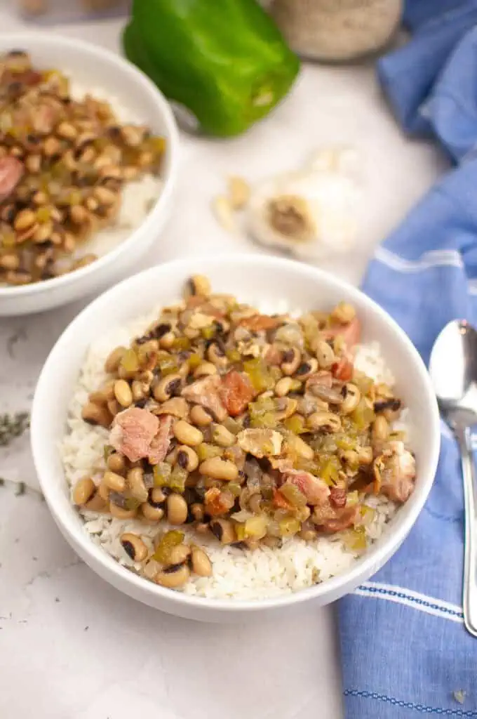 A bowl of rice topped with black-eyed peas, diced ham, and vegetables sits on a marble surface next to a spoon, a green bell pepper, garlic, and a blue napkin.
