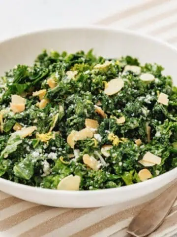 A white bowl of kale salad with lemon sits on a striped placemat beside a fork.