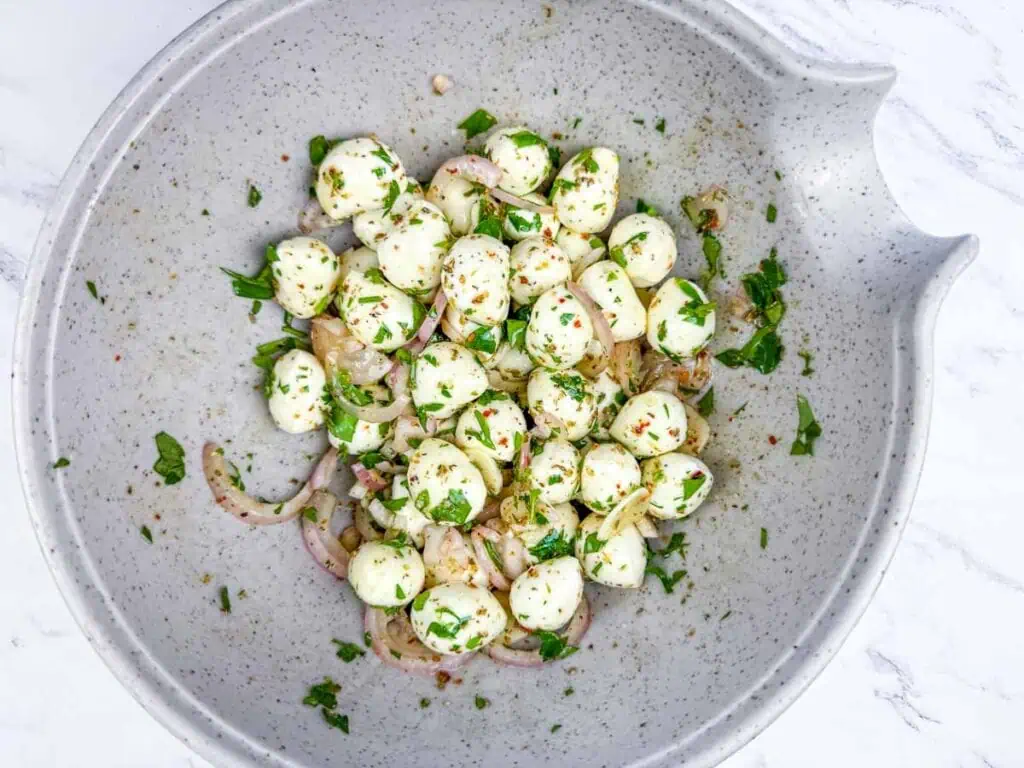A speckled bowl filled with mozzarella balls, sliced red onions, chopped herbs, and seasoning on a white marble surface.