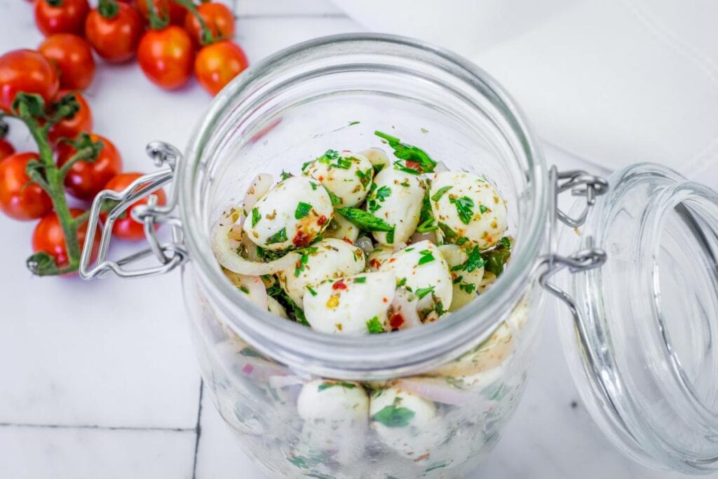 A glass jar filled with marinated mozzarella balls, onion slices, and herbs sits on a marble surface next to a vine of cherry tomatoes.