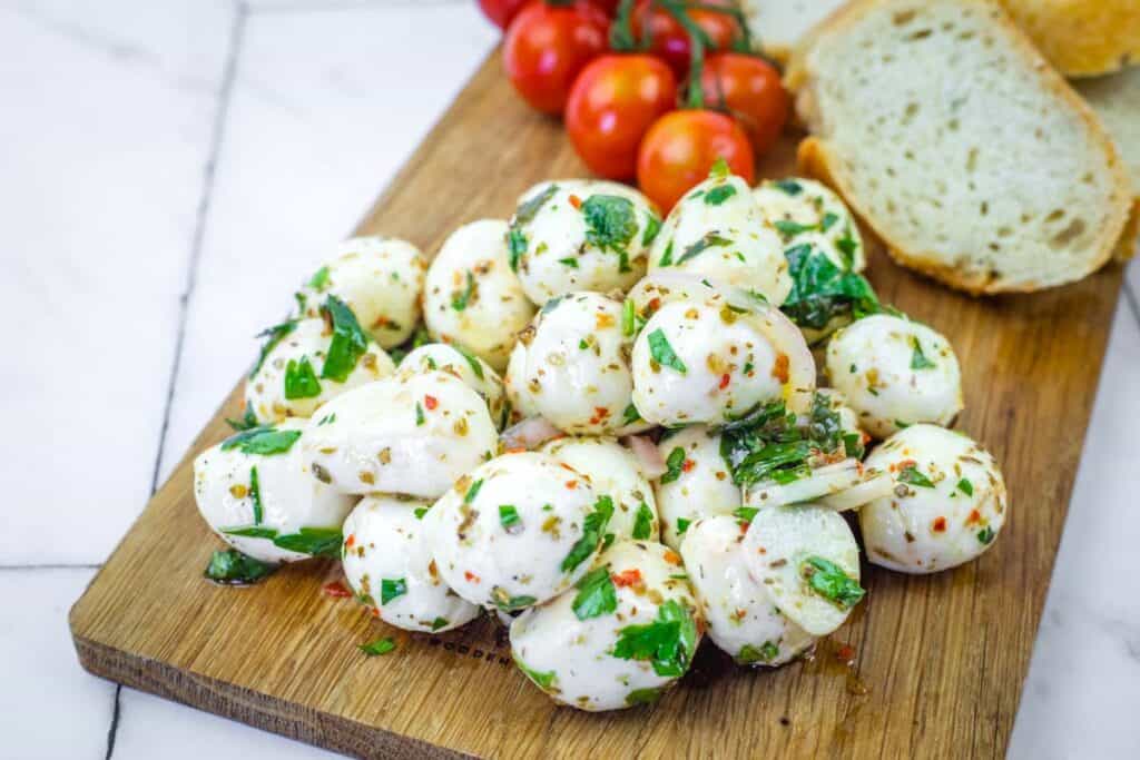 A pile of marinated mozzarella balls garnished with herbs on a wooden board, with cherry tomatoes and slices of bread in the background.