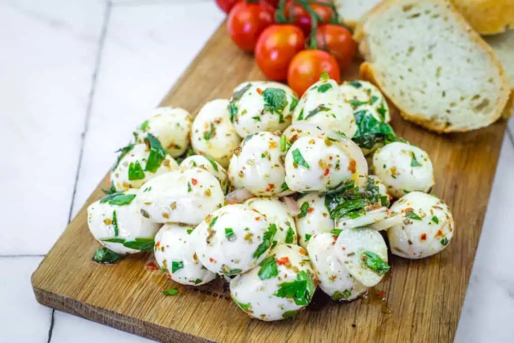 A pile of marinated mozzarella balls garnished with herbs on a wooden board, with cherry tomatoes and slices of bread in the background.