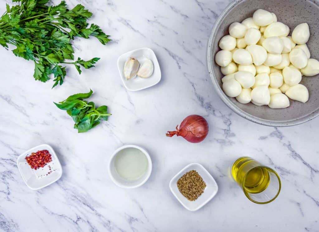 Flat lay of parsley, basil, garlic, shallot, red pepper flakes, vinegar, oregano, olive oil, and a bowl of peeled pearl onions on a marble surface.