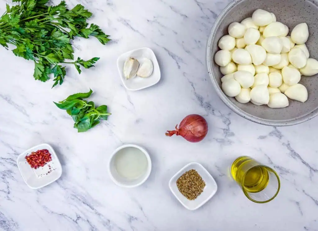 Flat lay of parsley, basil, garlic, shallot, red pepper flakes, vinegar, oregano, olive oil, and a bowl of peeled pearl onions on a marble surface.
