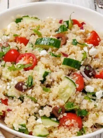 A bowl of Easy Mediterranean Quinoa Salad on a wooden surface with a striped cloth nearby.