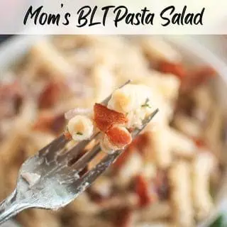 A close-up shot of a fork lifting pasta and bacon, with a blurry bowl of pasta salad in the background.