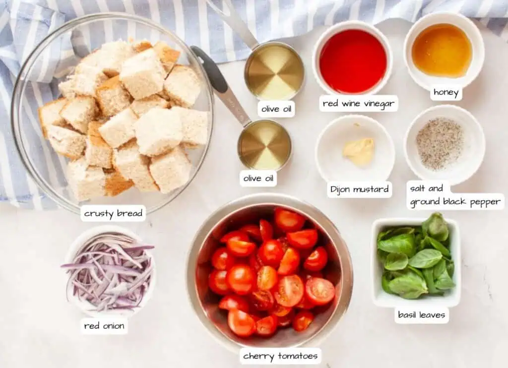 Overhead shot of panzanella salad ingredients, including bread cubes, tomatoes, red onion, basil, and dressing components, arranged on a light surface.