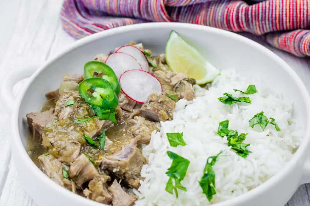 A white bowl filled with chile verde pork stew and white rice, garnished with sliced radishes, jalapenos, lime, and cilantro. A colorful cloth is in the background.