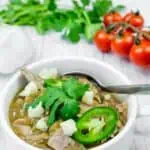 A white bowl filled with green pork chili, topped with cheese, jalapenos, and cilantro, with a spoon inside. Garlic, cilantro, and tomatoes are in the background.