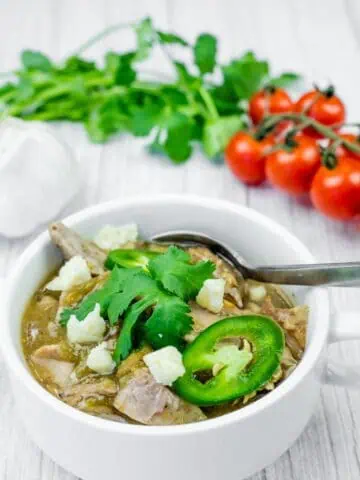 A white bowl filled with green pork chili, topped with cheese, jalapenos, and cilantro, with a spoon inside. Garlic, cilantro, and tomatoes are in the background.
