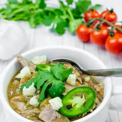 A white bowl filled with green pork chili, topped with cheese, jalapenos, and cilantro, with a spoon inside. Garlic, cilantro, and tomatoes are in the background.