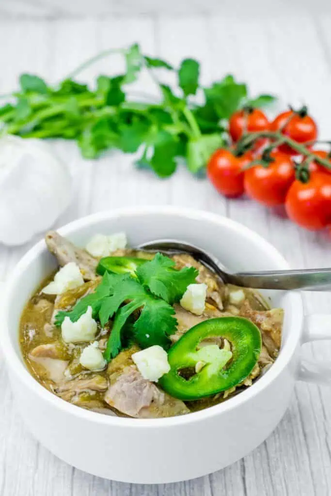 A white bowl filled with green pork chili, topped with cheese, jalapenos, and cilantro, with a spoon inside. Garlic, cilantro, and tomatoes are in the background.