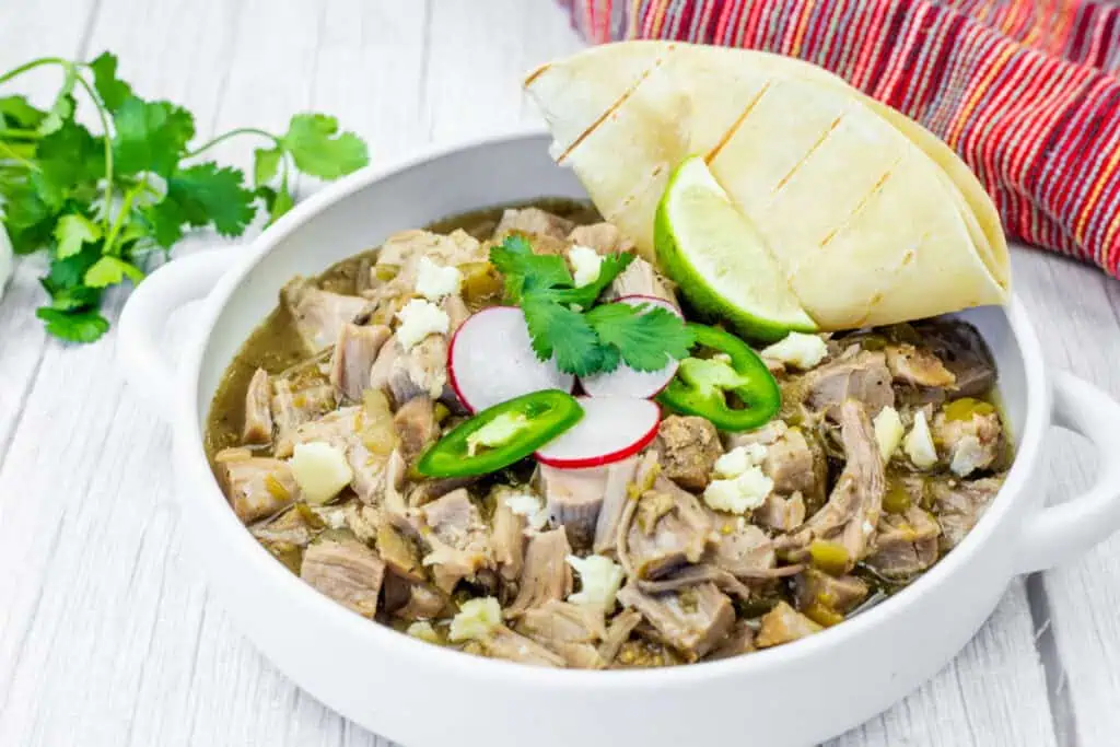 A white bowl of Slow Cooker Pork Chile Verde, featuring visible chunks of meat, radish slices, jalapeno slices, cilantro, and cheese, sits on a white wooden table. A folded tortilla and lime wedge are placed beside the bowl. A head of garlic and cilantro sprigs are in the background, along with a red and white striped cloth.