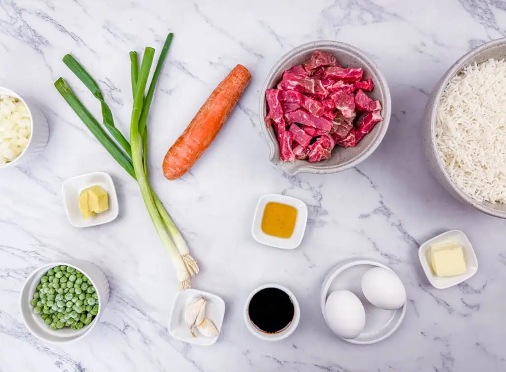 Rice, sliced steak, carrot, oil, eggs, butter, soy sauce, garlic, peeled ginger, sliced onion, green onion, and peas on a marbled white-gray surface
