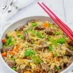 Steak fried rice in a white bowl with handles, red chopsticks on top and folded white napkin on background.