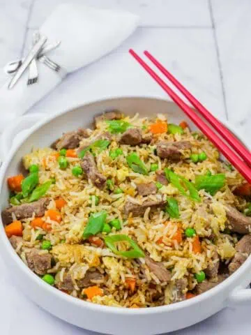 Steak fried rice in a white bowl with handles, red chopsticks on top and folded white napkin on background.