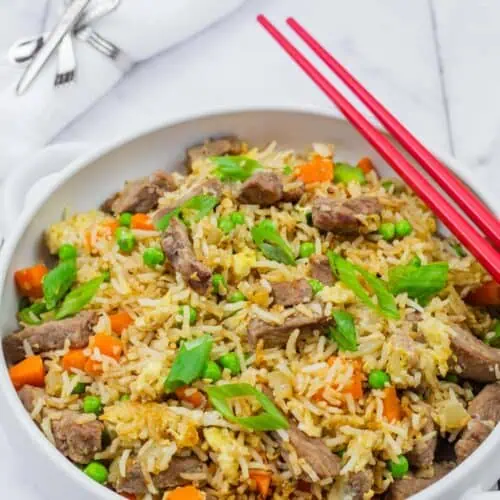Steak fried rice in a white bowl with handles, red chopsticks on top and folded white napkin on background.