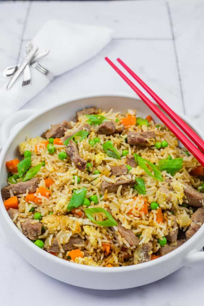 Steak fried rice in a white bowl with handles, red chopsticks on top and folded white napkin on background.