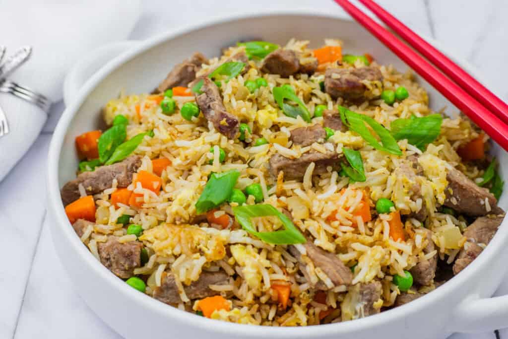 steak fried rice in a white bowl with handles and red chopsticks on top.