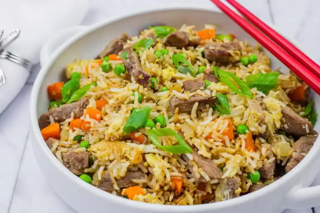 steak fried rice in a white bowl with handles and red chopsticks on top.
