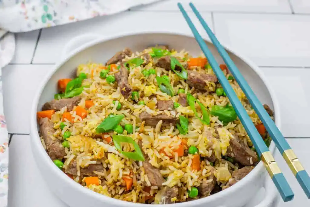 A bowl of steak fried rice served with blue chopsticks on a white tiled surface.