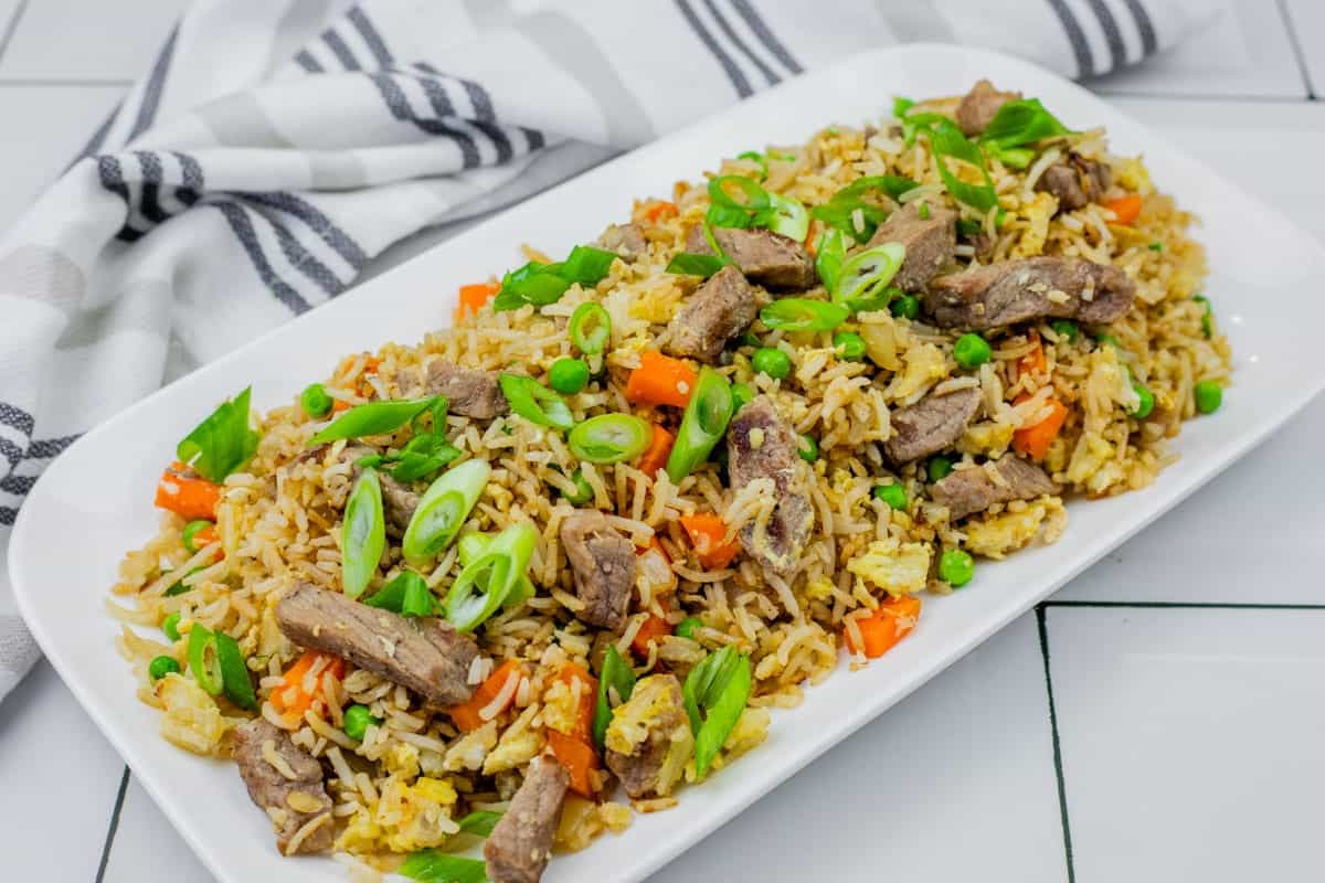 Steak fried rice on a white platter on white tiles with striped cloth on the side.