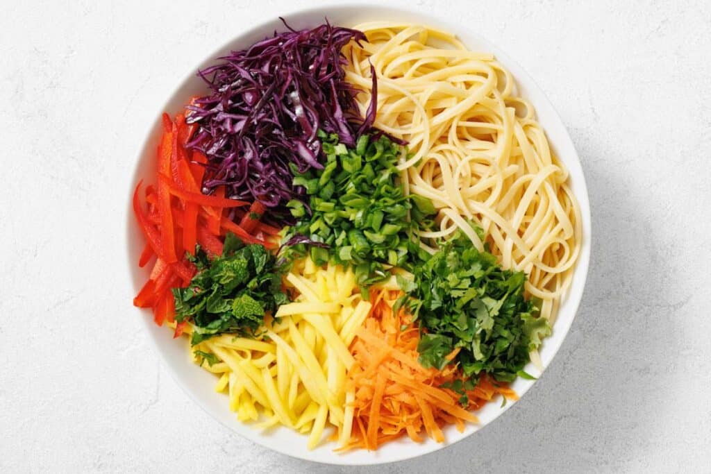 A white bowl filled with sections of cooked noodles, shredded red cabbage, sliced red bell peppers, chopped green onions, mint, mango, cilantro, and grated carrot.