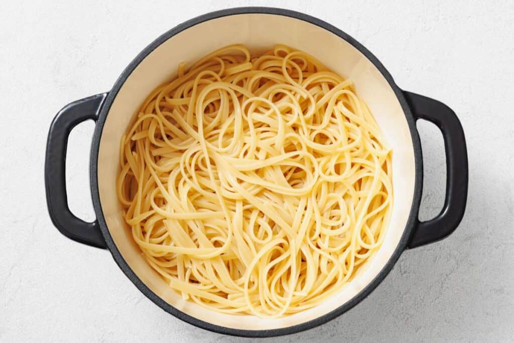 A black-handled pot filled with cooked plain spaghetti noodles on a light-colored surface.