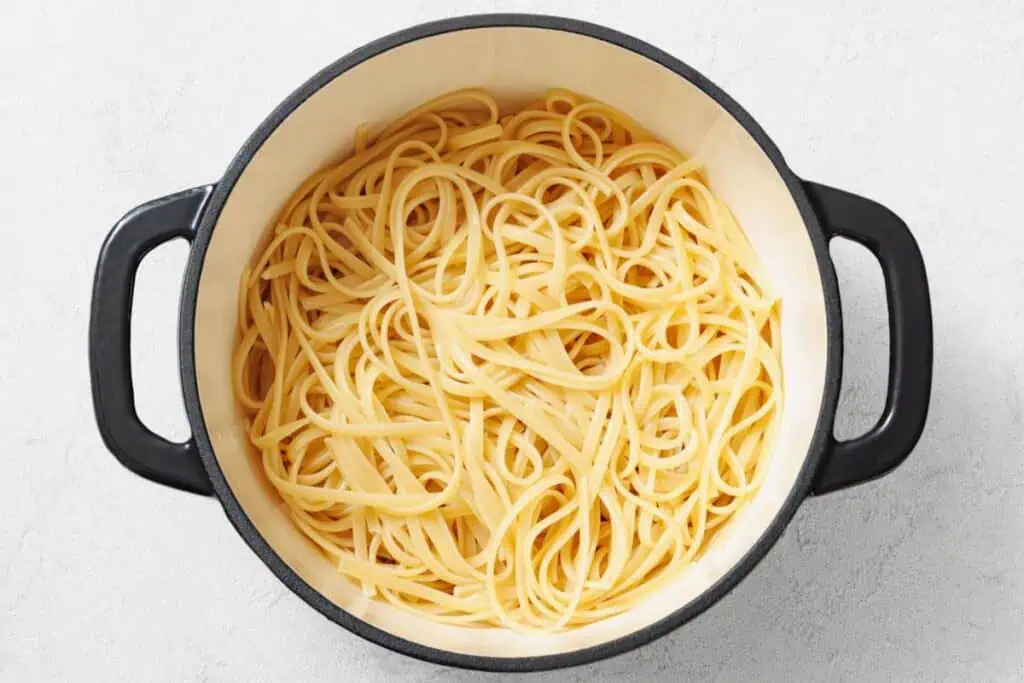 A black-handled pot filled with cooked plain spaghetti noodles on a light-colored surface.