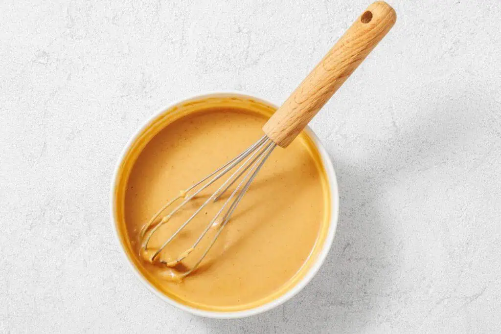 A metal whisk with a wooden handle rests in a bowl of creamy, light brown batter or sauce on a light gray surface.