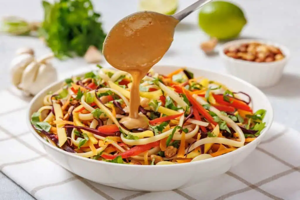 A spoon pours creamy dressing over a bowl of colorful vegetable noodles, garnished with herbs and nuts, on a checkered napkin.